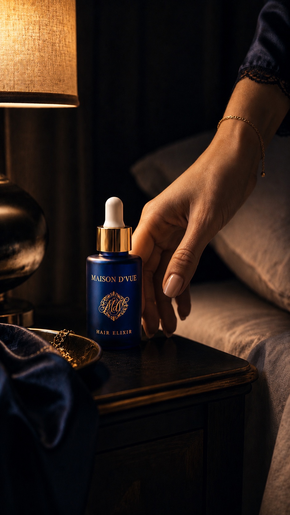 The MAISON D'VUE Hair Elixir on a bedside table beside a lit lamp, a hand reaching for it